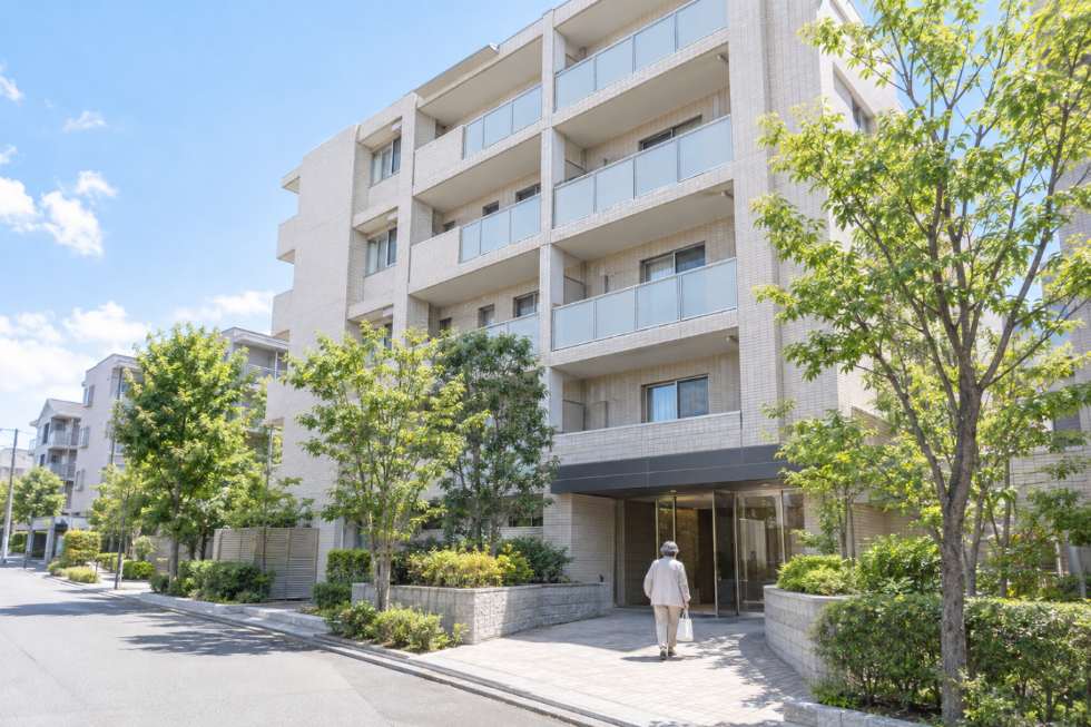 晴れた日の中規模賃貸マンションと、買い物袋を提げて帰宅する高齢の入居者の写真イメージ